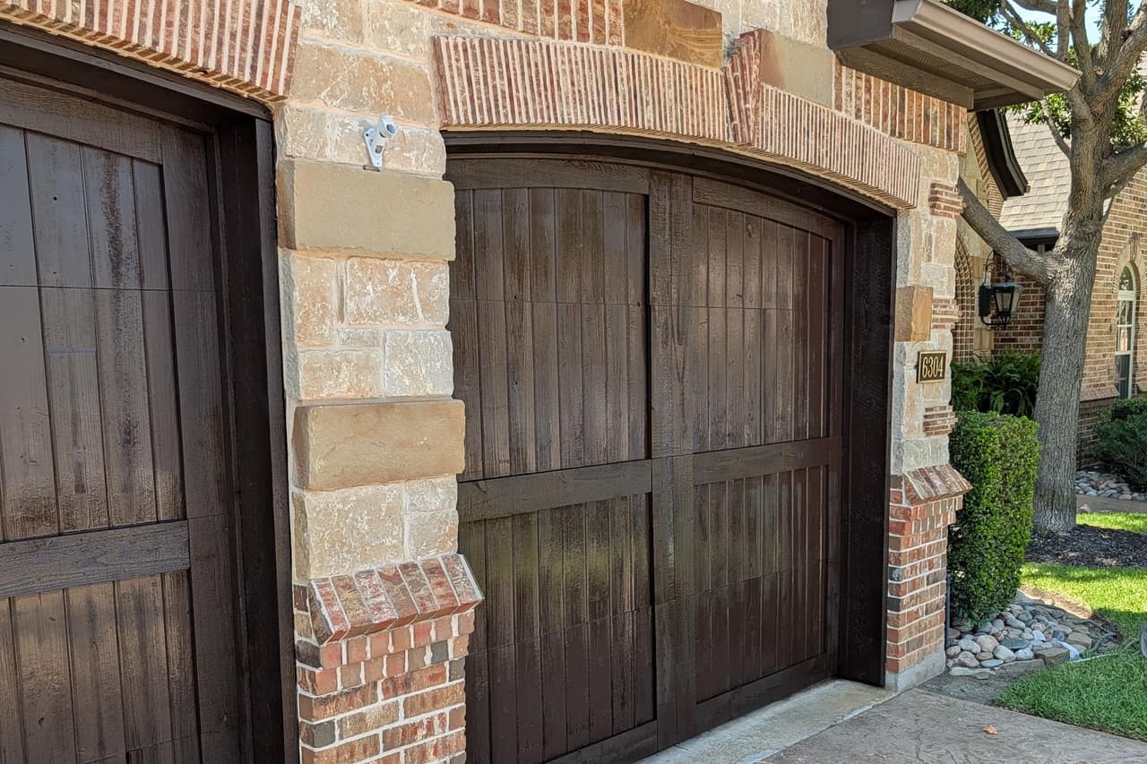 Two arched wood garage doors freshly stained a deep walnut, framed by stone and brick